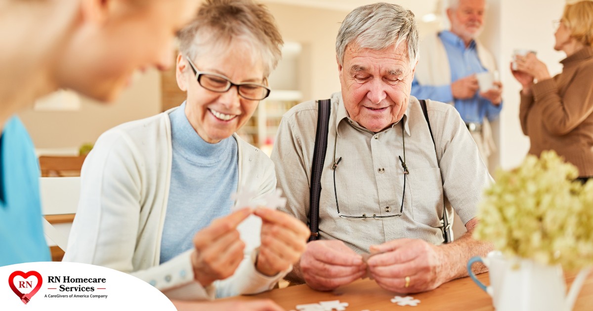 An older couple works on a puzzle with a caregiver, representing the kind of activity that helps those with dementia and also representing Alzheimer’s and Brain Awareness Month.