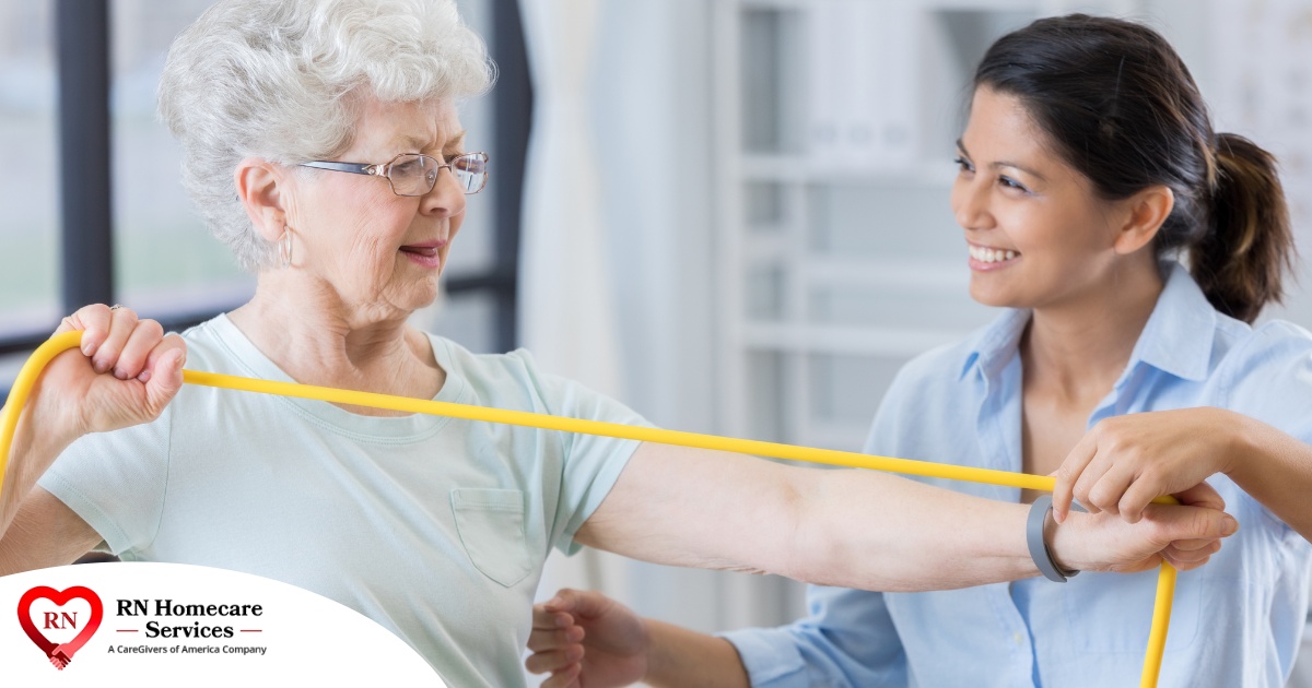 A woman helps an older woman exercise with a resistance band, showing how in-home physical therapy can help.